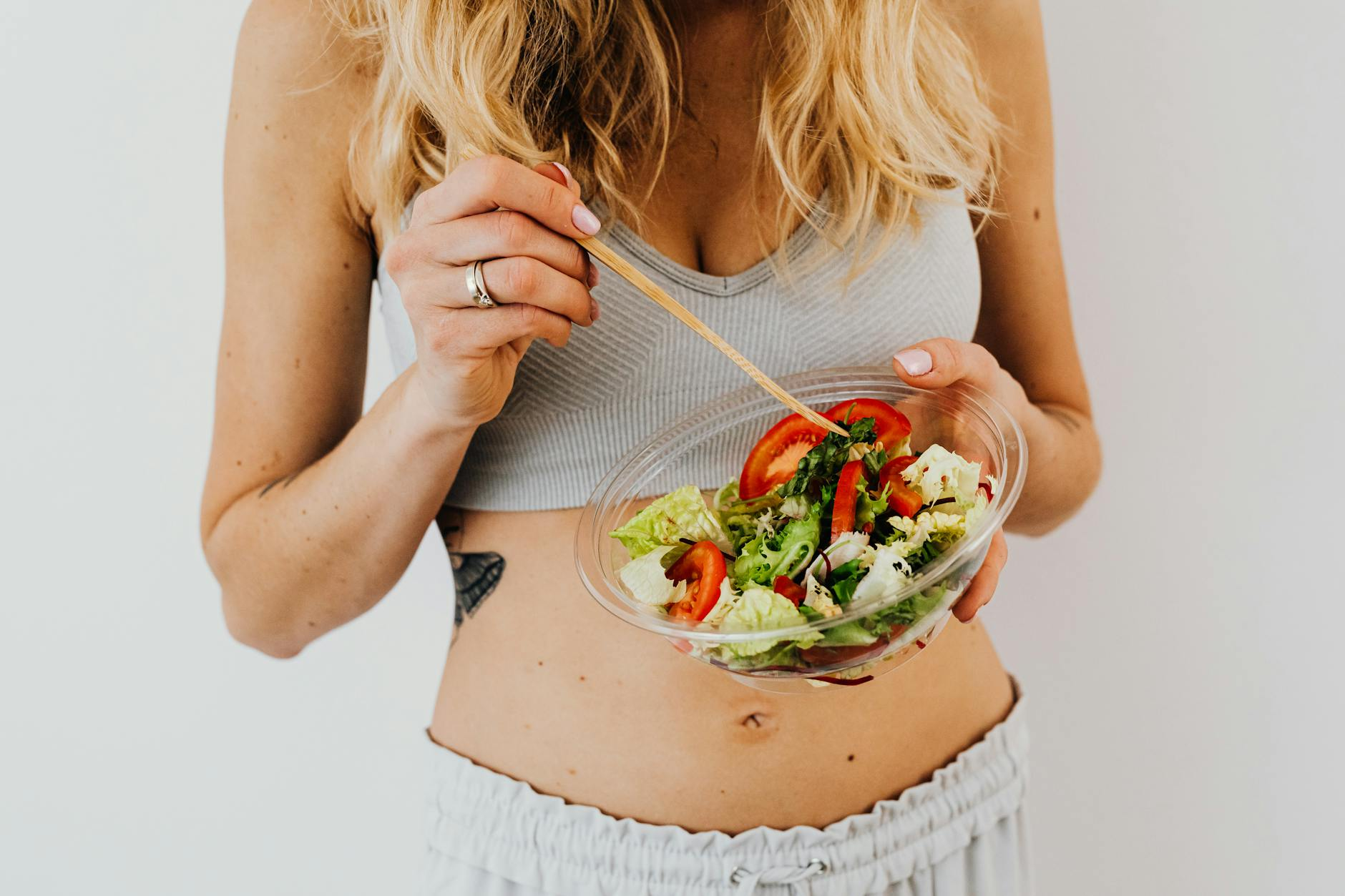 Eine schwangere Frau mit freundlichem Lächeln sitzt an einem Holztisch in einer hellen Küche und betrachtet eine Auswahl an frischem Obst, Gemüse und Vollkornprodukten, die eine gesunde Ernährung in der Schwangerschaft darstellen.