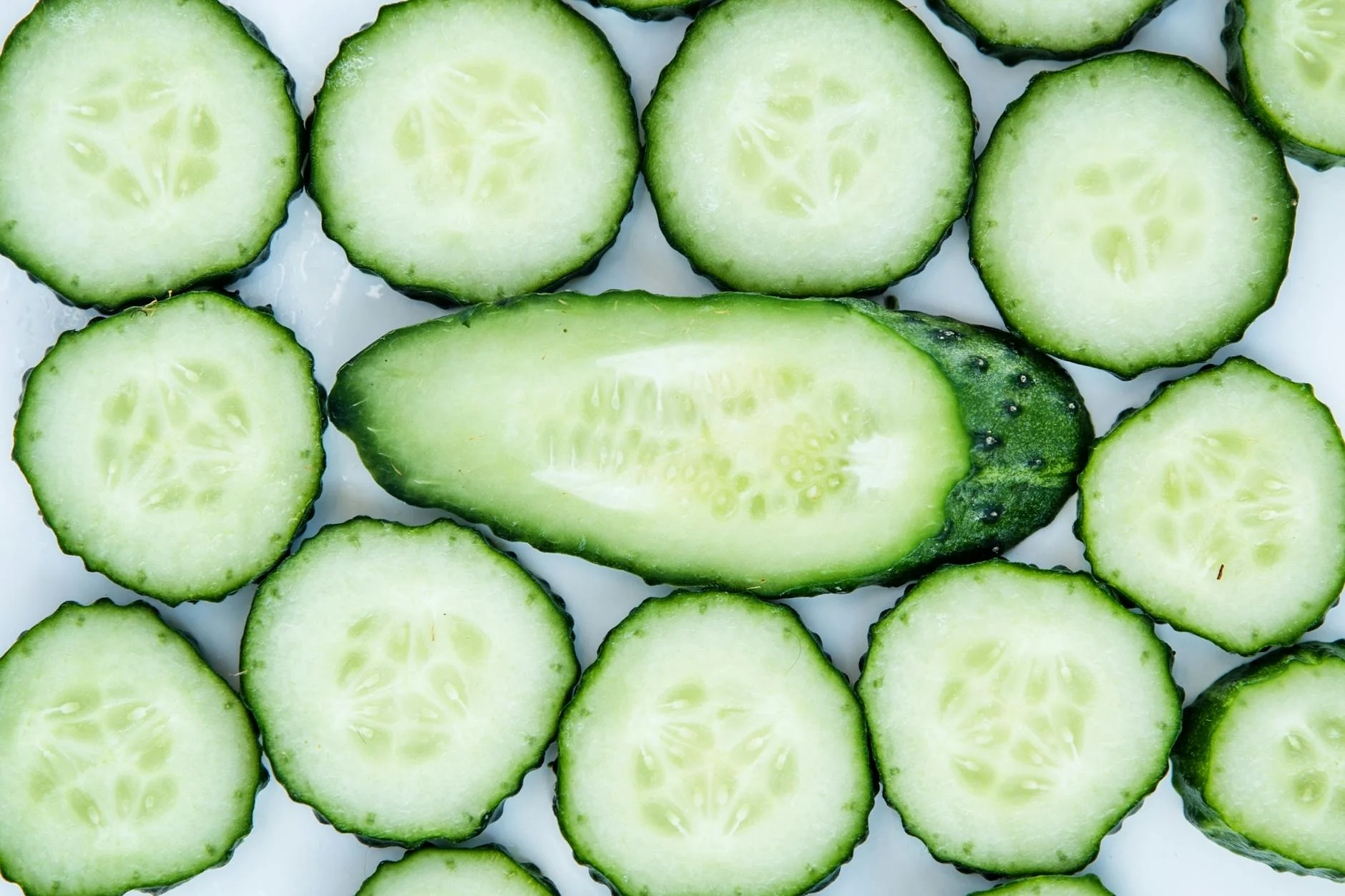 Nahaufnahme einer Frau mit sichtbaren dunklen Augenringen und leichten Tränensäcken, daneben liegen Gurkenscheiben und eine Wasserflasche als natürliche Hilfsmittel.