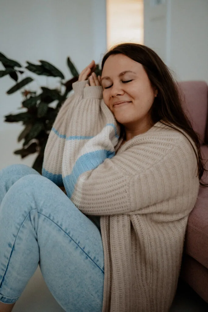 Johanna smiling warmly on pink sofa, embodying the radiant health and ease she helps clients achieve.